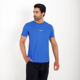 Modelo usando Camisetas Poliamida UV30 azul em fundo branco, com uma mão no bolso.